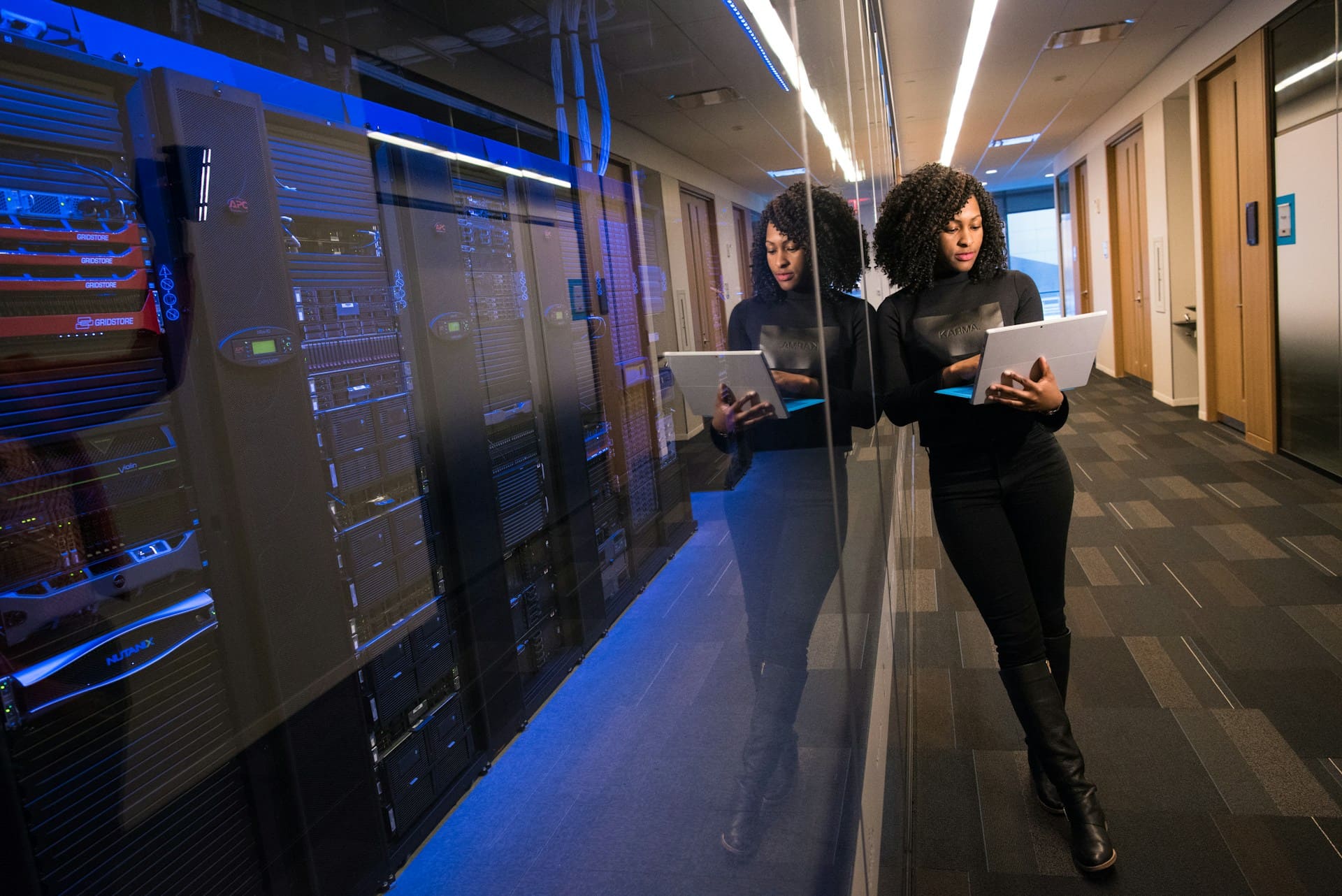 Woman working on a laptop in a hallway beside a glass wall reflecting rows of data servers